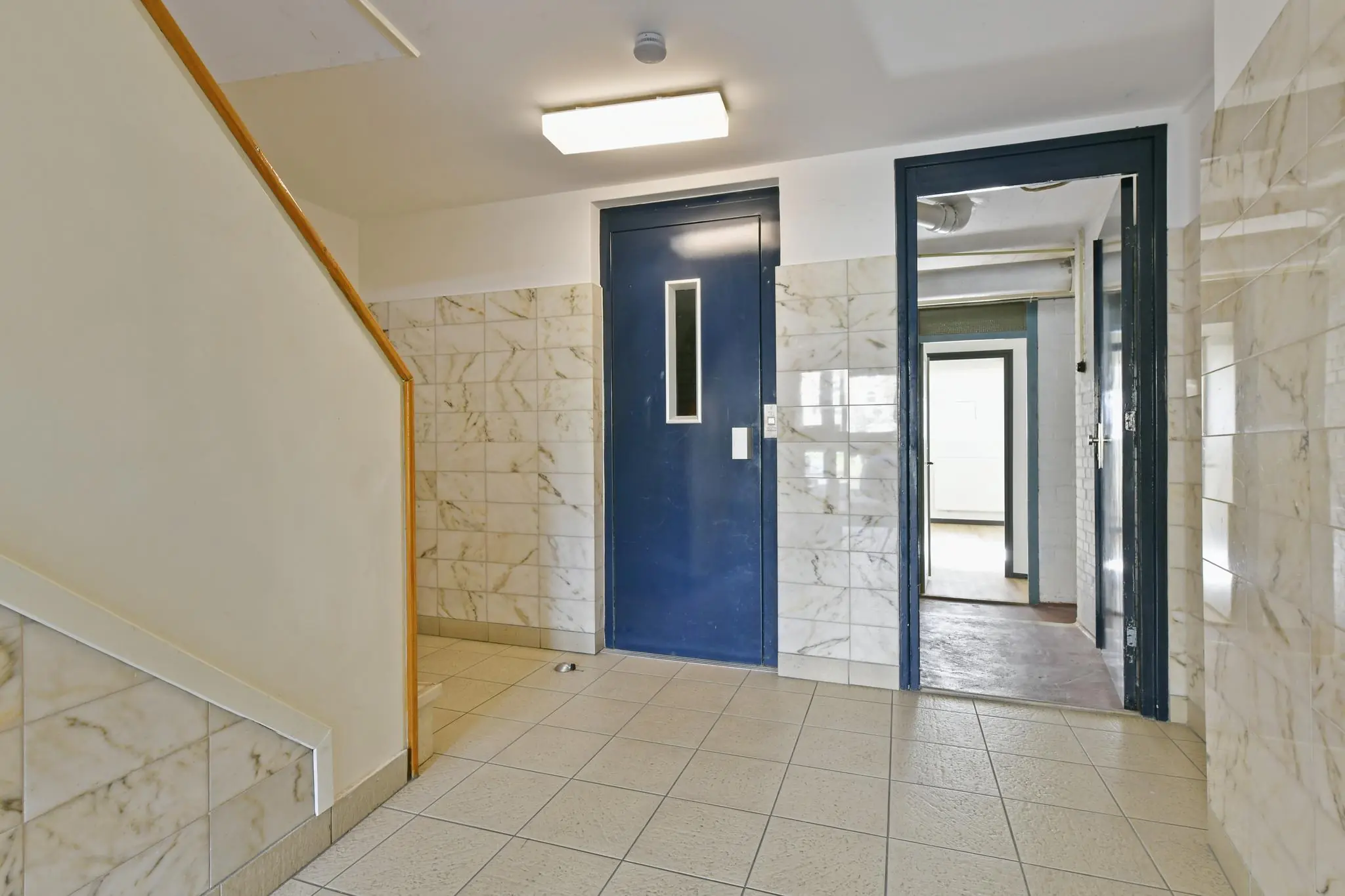 Trappenhuis met marmeren wandtegels, een blauwe liftdeur en een open doorgang in een gebouw aan de Cornelis Dopperkade.